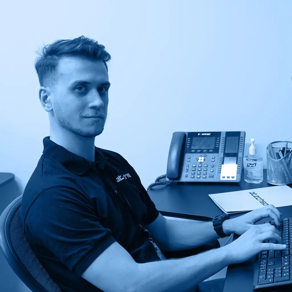 Young male technician in a black shirt working at a computer desk with a phone in view.