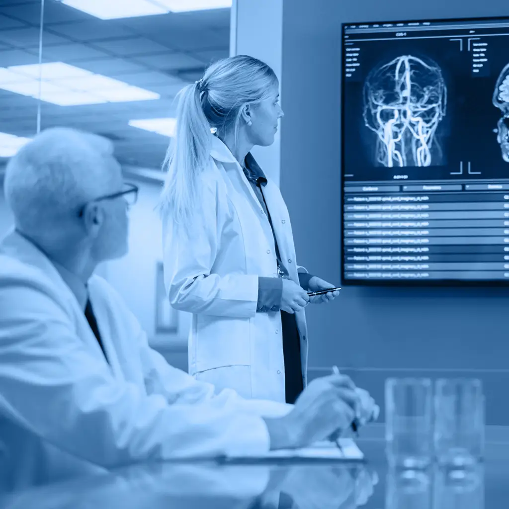 Female medical professional presenting a brain scan on monitors during a clinical meeting with colleagues.=