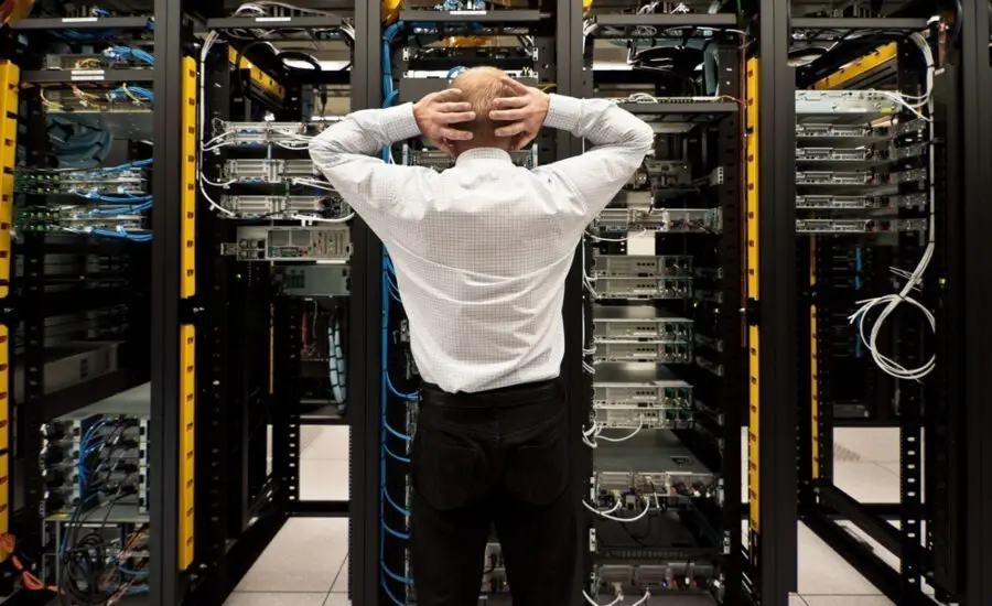 Frustrated IT professional in a server room with hands on head, standing between racks of network equipment, reflecting challenges in data center management or technical difficulties.