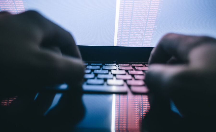 Close-up of hands typing on a backlit laptop keyboard with reflections of red and blue code on the screen, suggesting cybersecurity or hacking activity